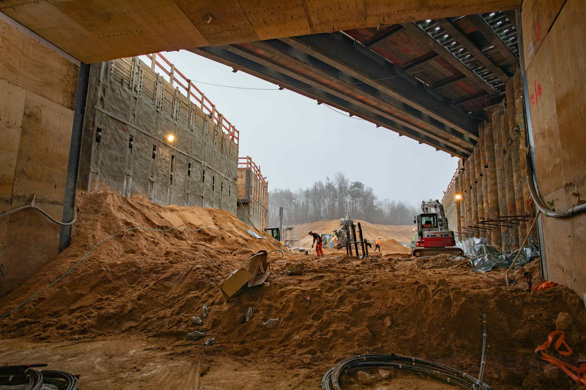 Blick aus dem Tunnel auf Mengen von Erde, einem Bagger und Bauarbeitern, im Hintergrund Bäume